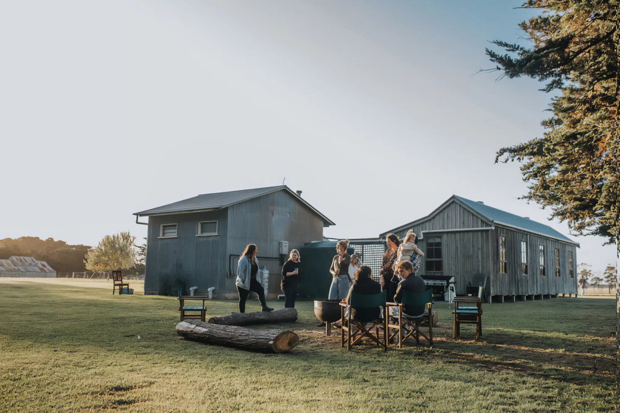 Barooma Homestead - picturesque pastorial gathering of friends having a drink at sunset while standing outside a series of rustic farm buildings